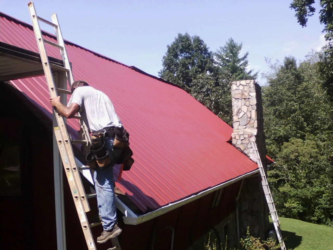 Roofer on a ladder beside a red metal roof and stone chimney — Soffit & Fascia Repair by Roofing Brothers in Barbourville